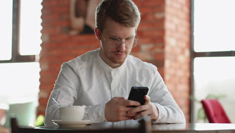 Ein-Erwachsener-Attraktiver-Mann-Benutzt-Ein-Smartphone,-Sitzt-Allein-Im-Café-Und-Wartet-Auf-Jemanden-Und-Chattet-Online